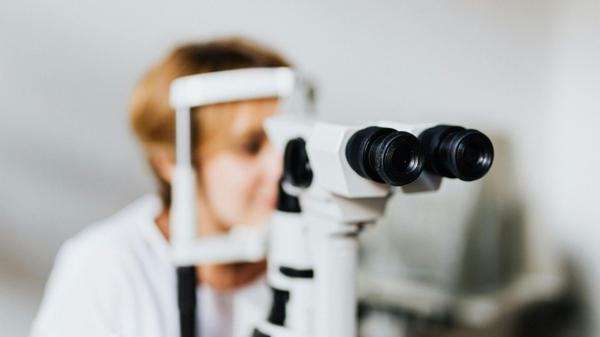 A high-end perspective focusing through the eyepieces of an optometry slit lamp microscope, with an optometrist blurred in the background.